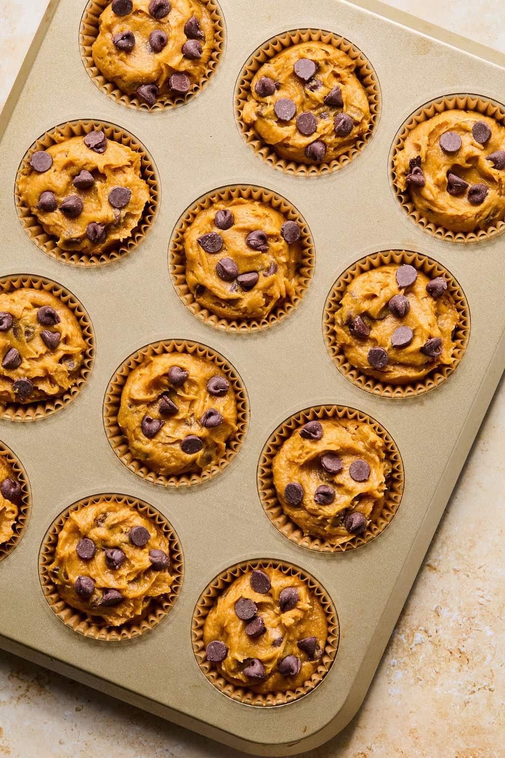 A muffin tin filled with unbaked gluten-free pumpkin muffins batter, each lined with a paper cup and topped with chocolate chips, sits on a light-colored countertop.