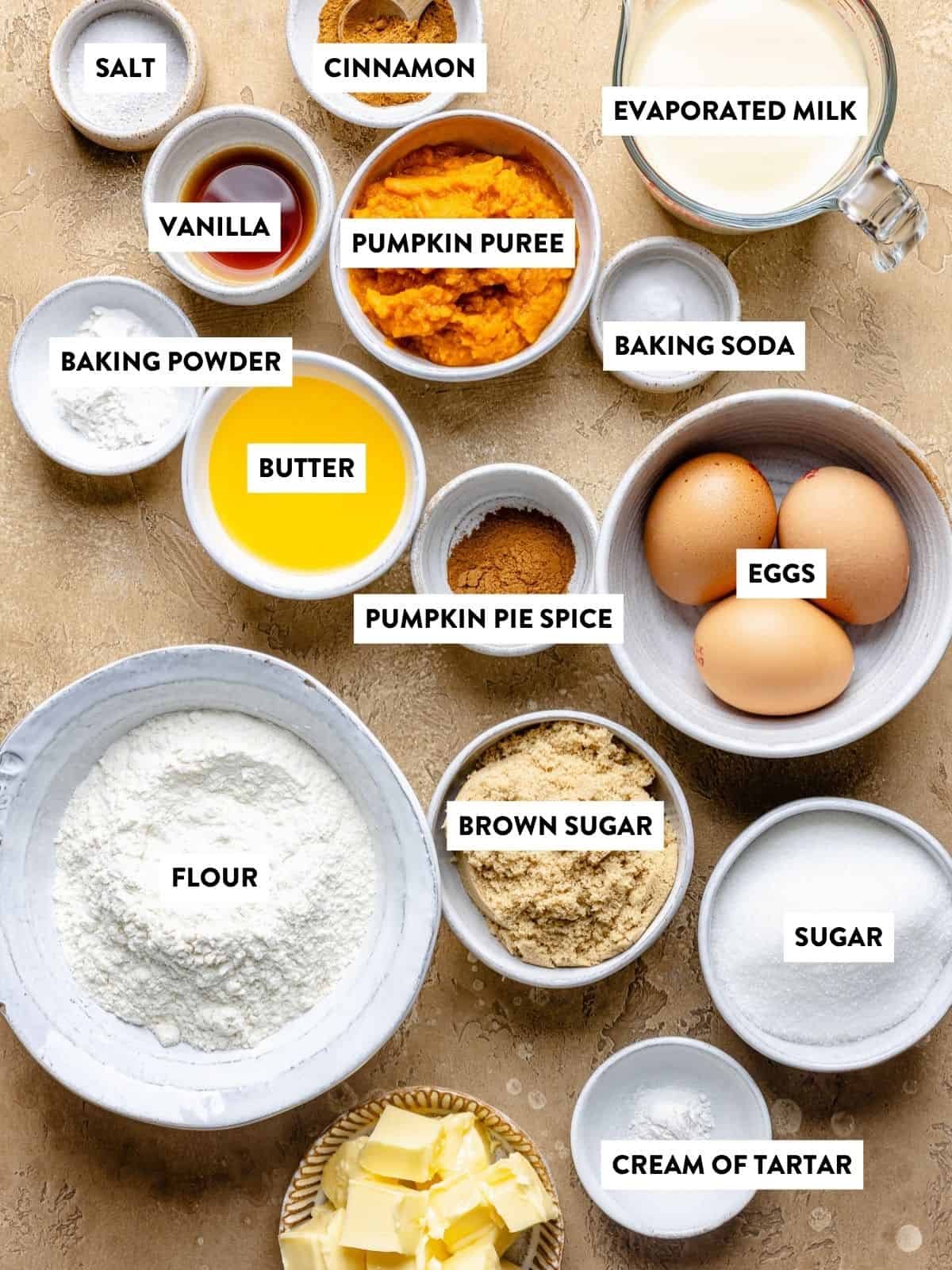 Various baking ingredients labeled in bowls and dishes, including flour, brown sugar, sugar, eggs, butter, pumpkin puree, evaporated milk, vanilla, spices, cream of tartar, baking powder, baking soda, and salt—everything you need to create a delightful snickerdoodle cobbler.