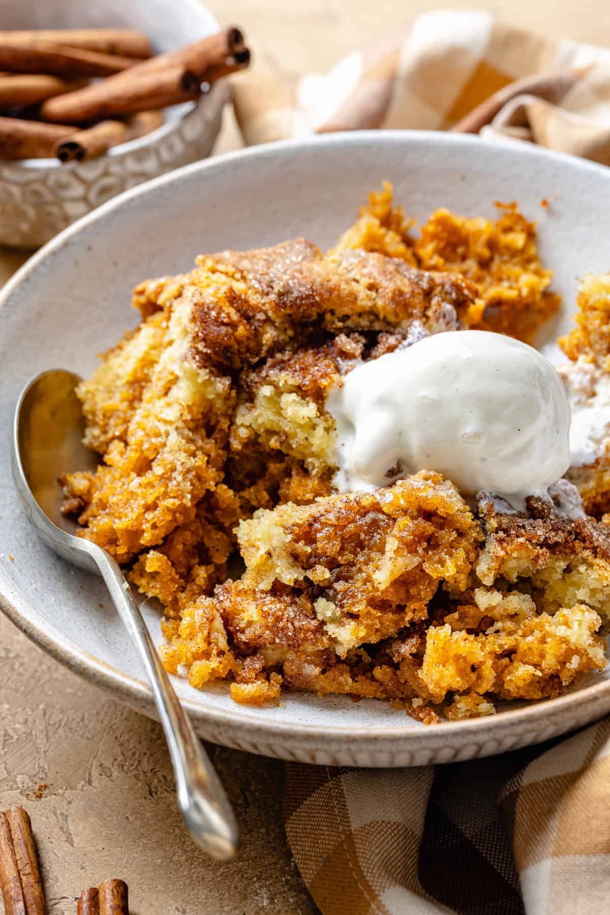 A bowl of pumpkin cobbler topped with a scoop of vanilla ice cream and set next to a spoon and a dish of cinnamon sticks in the background.
