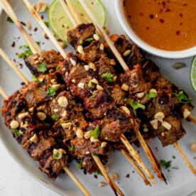 grilled beef satay on a white serving plate topped with peanuts and cilantro with a side of peanut sauce