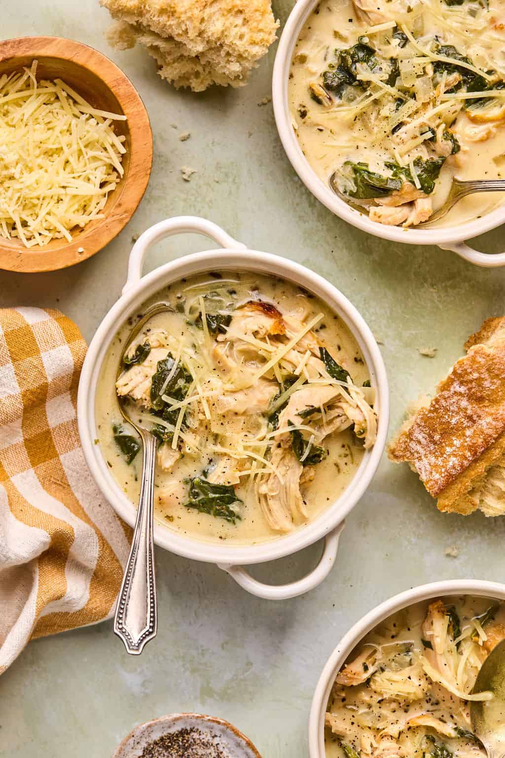 three bowls of chicken florentine soup on a counter with parmesan cheese and bread.