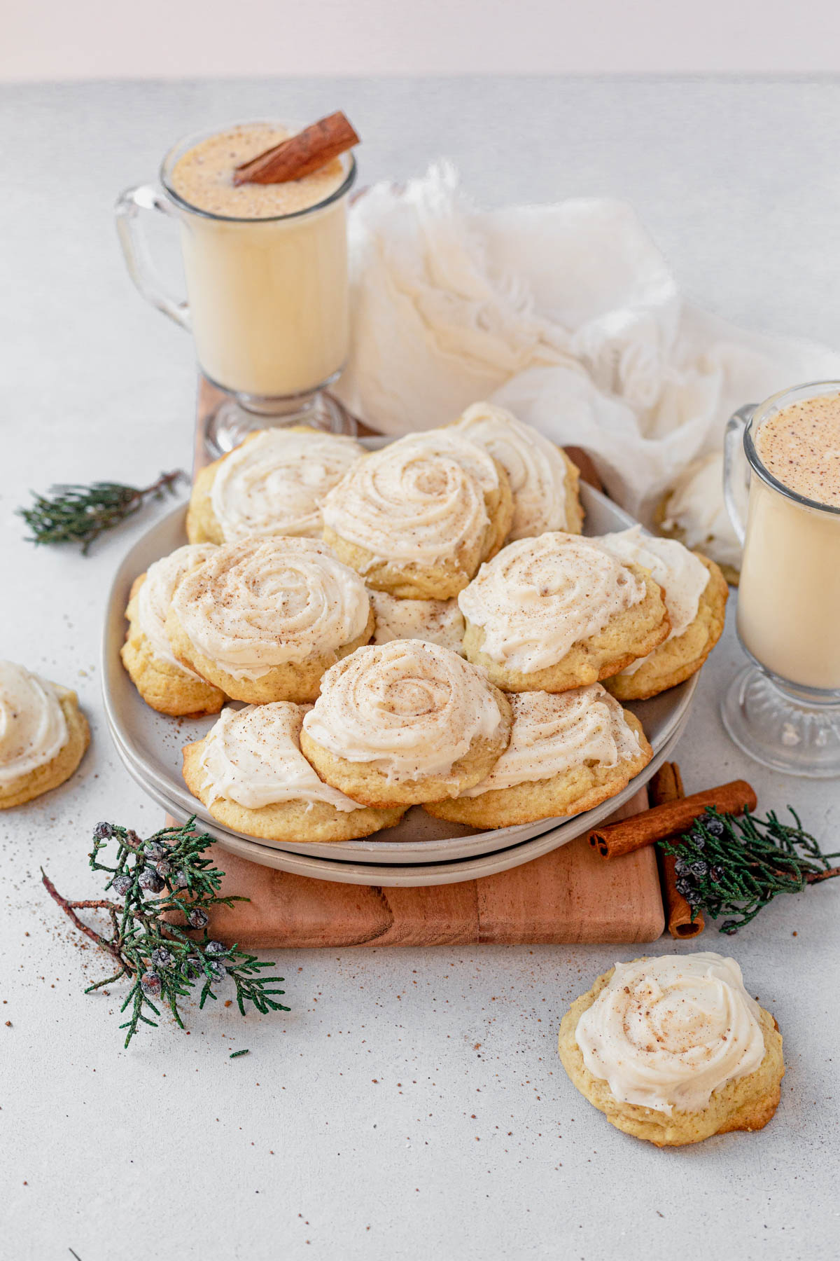 a plate of eggnog cookies surrounded by cups of eggnog and evergreen sprigs