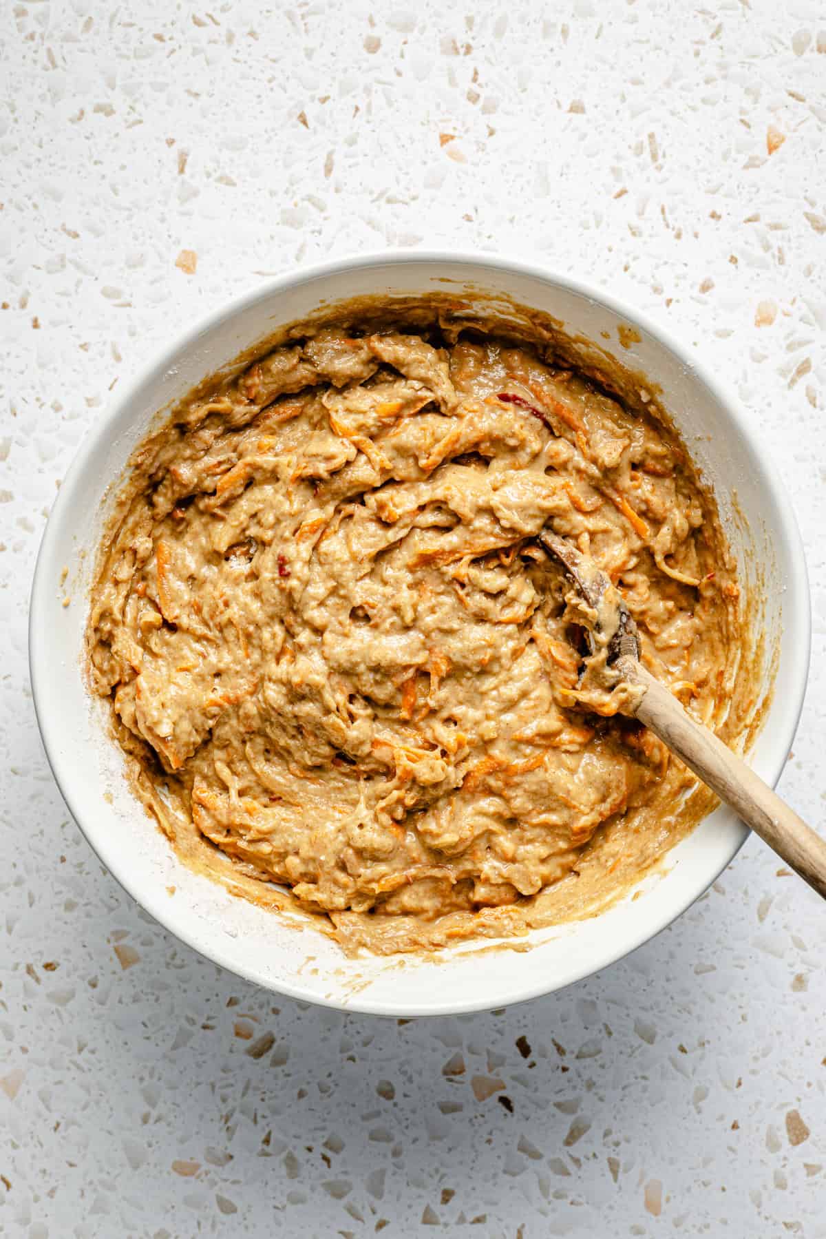 healthy morning glory muffin batter resting in a bowl on the counter with a wooden spoon resting in it.