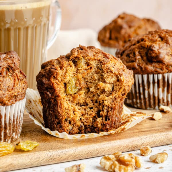 a healthy morning glroy muffin on a wood serving tray with the wrapper removed and a bite taken out surrounded by a latte and more muffins.