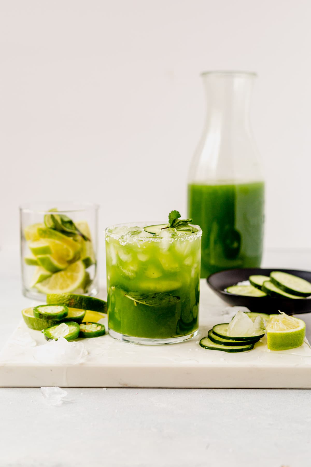 a fresh cucumber margarita in a glass with the pitcher of margaritas behind it