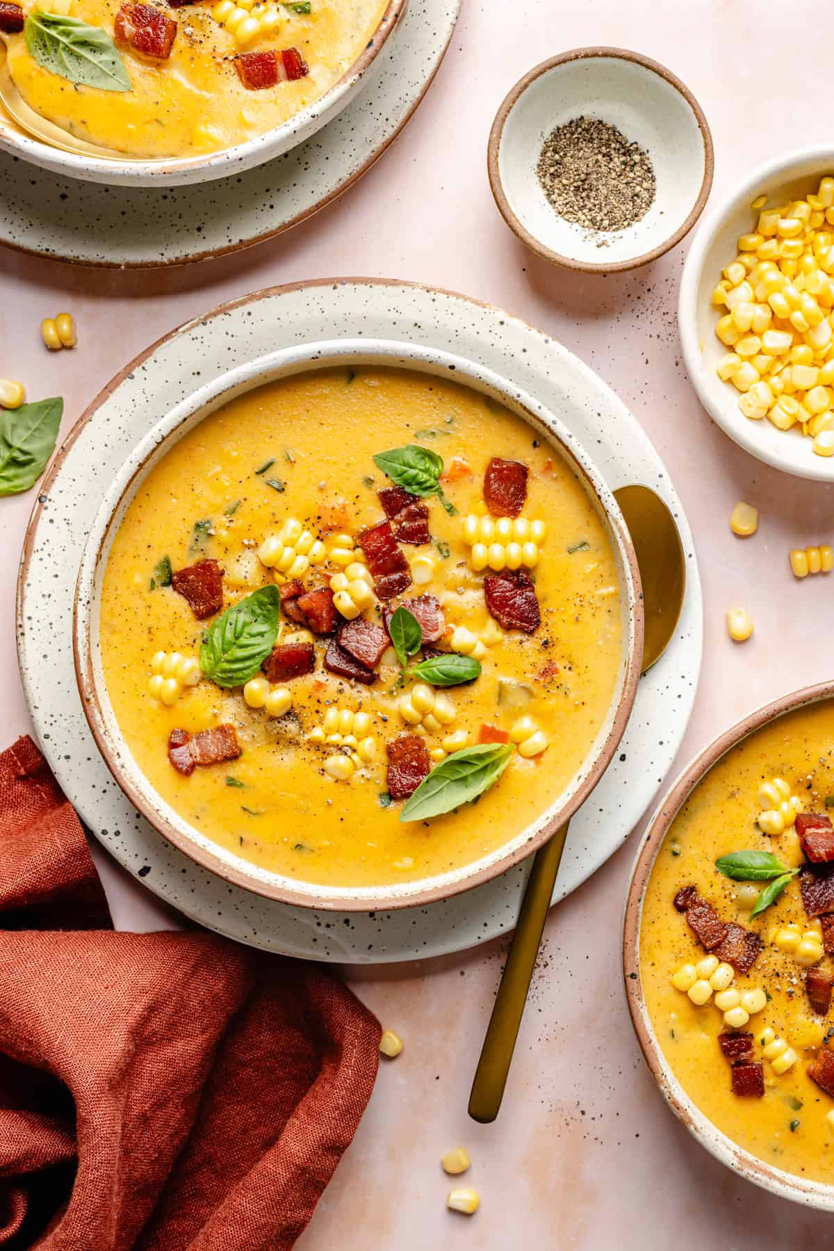 Three bowls of creamy instant pot corn chowder topped with bacon, corn kernels, and basil leaves, surrounded by a napkin, spoon, and small dishes of pepper and corn on a pink background.