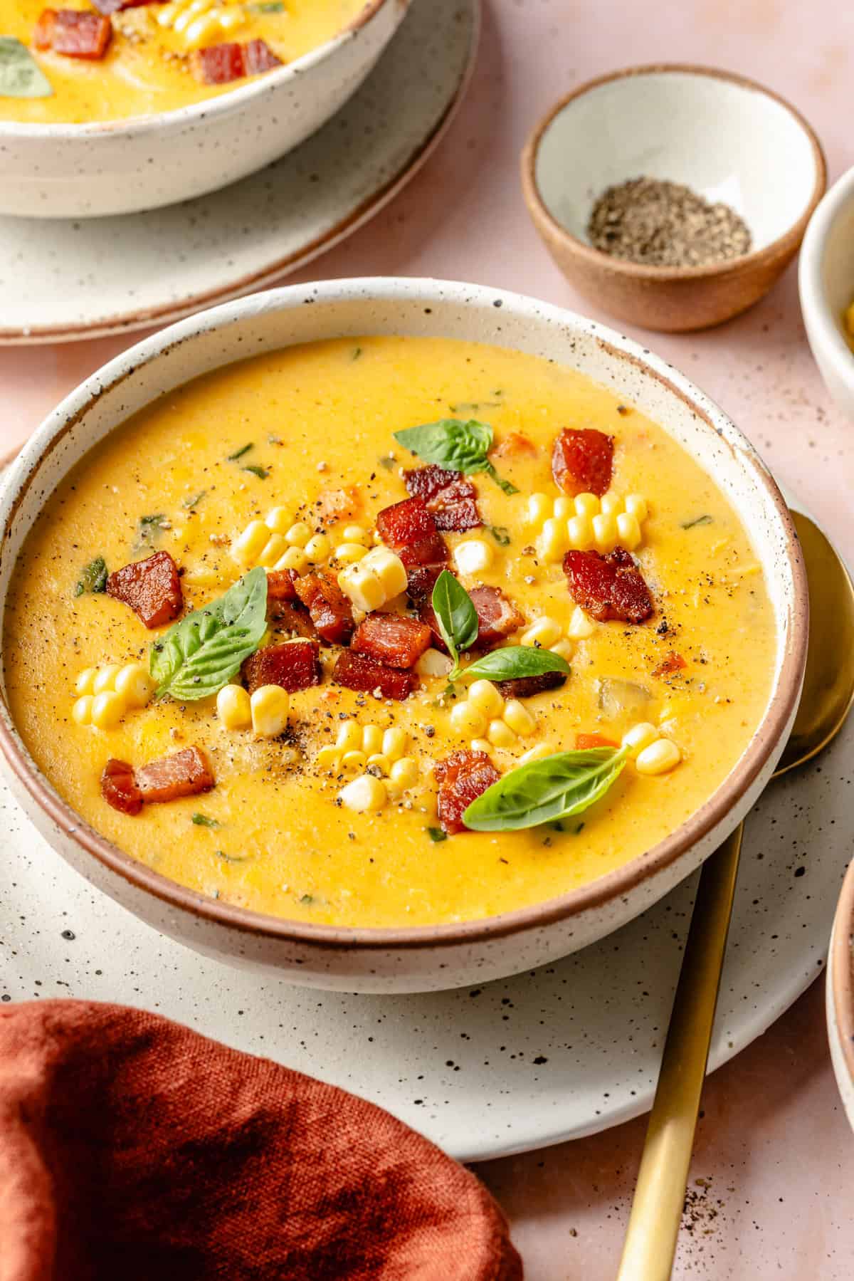 A bowl of creamy Instant Pot corn chowder topped with bacon, fresh corn kernels, basil leaves, and black pepper, served on a ceramic plate with a gold spoon and napkin nearby.