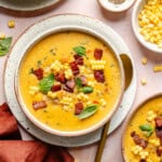 A bowl of creamy Instant Pot corn chowder topped with crispy bacon, corn kernels, and fresh basil leaves, with a gold spoon on the side and bowls of extra corn and pepper nearby.