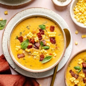 A bowl of creamy Instant Pot corn chowder topped with crispy bacon, corn kernels, and fresh basil leaves, with a gold spoon on the side and bowls of extra corn and pepper nearby.