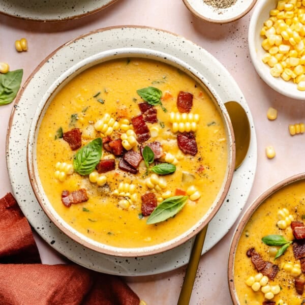 A bowl of creamy Instant Pot corn chowder topped with crispy bacon, corn kernels, and fresh basil leaves, with a gold spoon on the side and bowls of extra corn and pepper nearby.