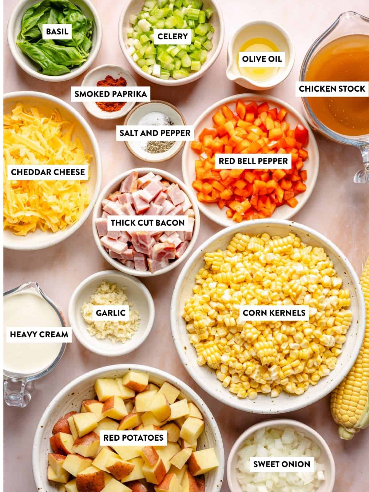 Assorted labeled ingredients in bowls for a recipe of instant pot corn chowder, including corn kernels, red potatoes, bacon, cheddar cheese, red bell pepper, celery, sweet onion, garlic, heavy cream, olive oil, chicken stock, basil, and spices.