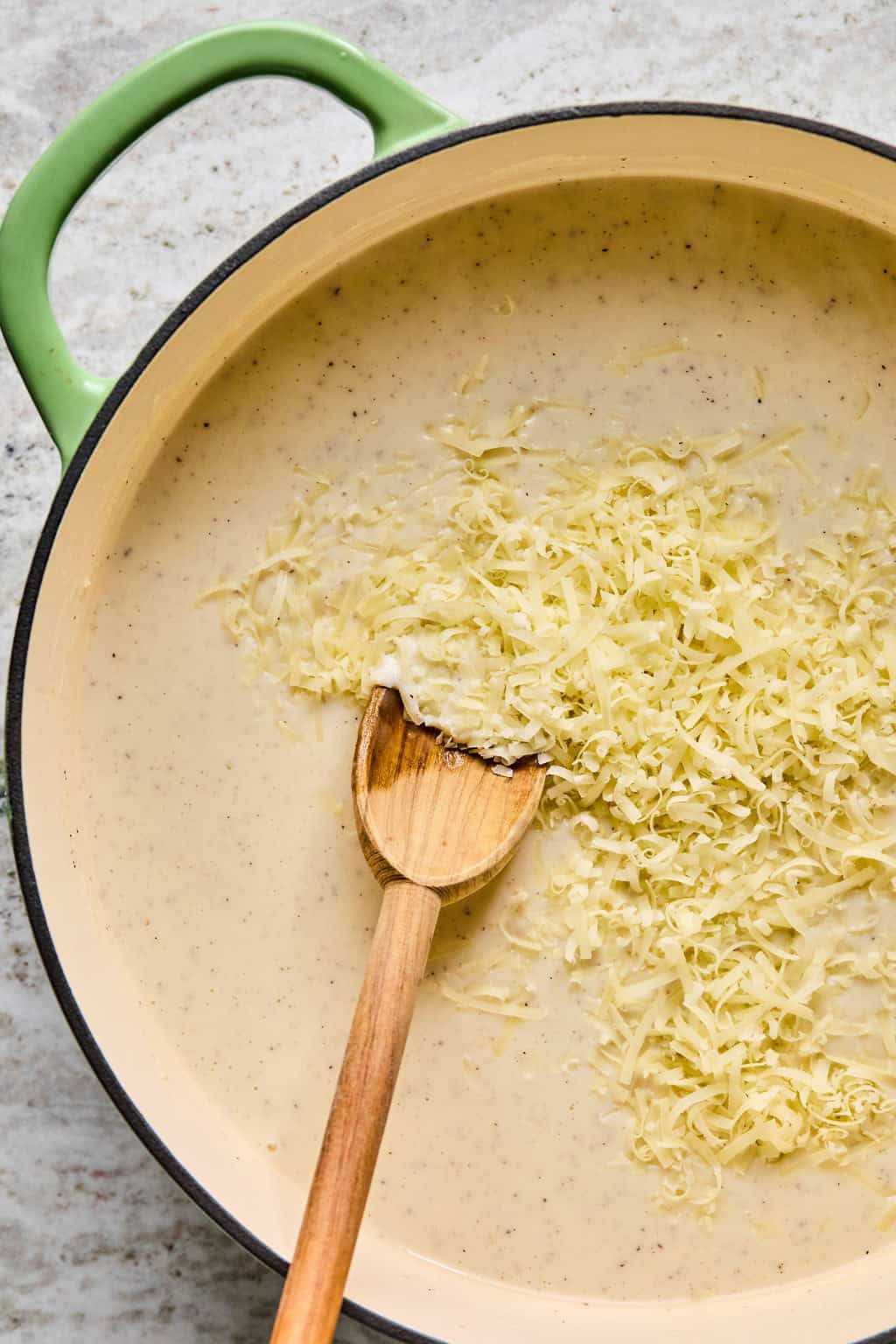 A wooden spoon stirs shredded cheese into a creamy white sauce for steak pasta in a large, round, light-colored pot with a green handle, set on a light countertop.