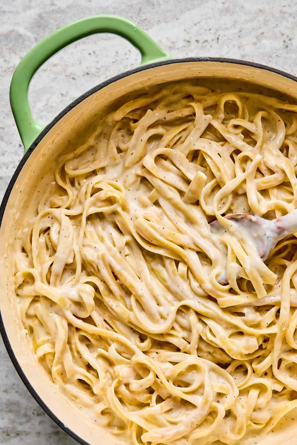 A pot with a green handle filled with creamy steak pasta Alfredo, being stirred with a wooden spoon on a light textured surface.