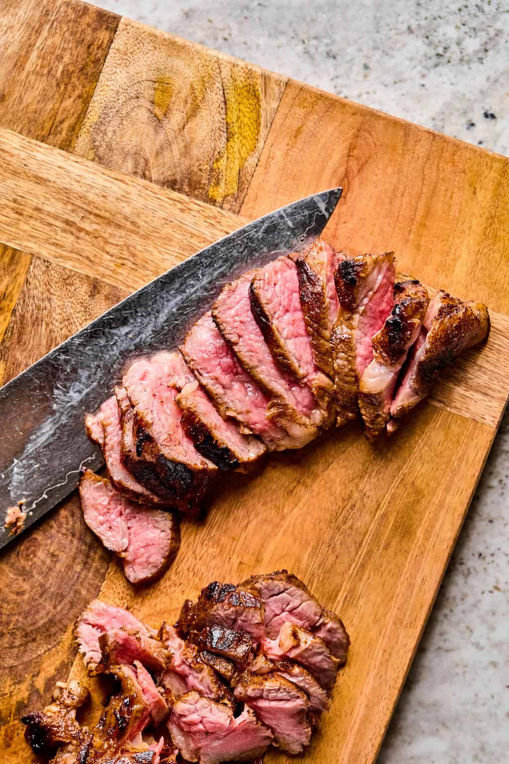 Sliced medium-rare steak on a wooden cutting board with a large knife beside it. The seared, brown crust and pink center pair perfectly with steak pasta, with savory juices visible on the board.