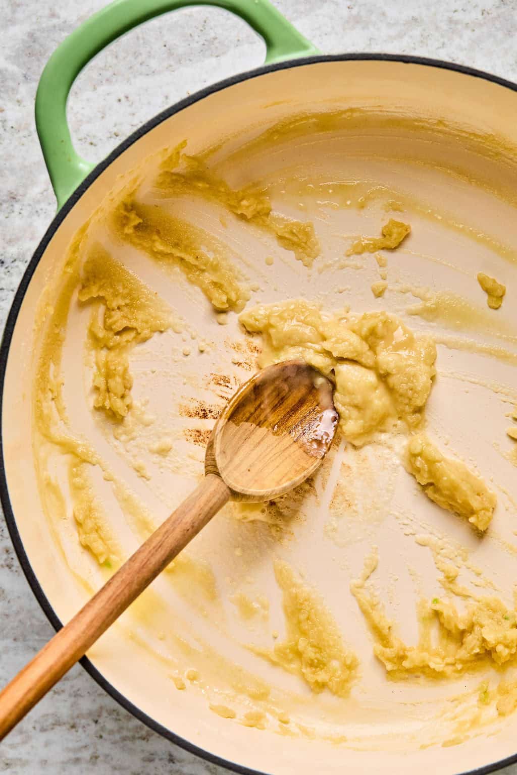 A wooden spoon stirs a thick, light-yellow roux in a large cream-colored pot with a green handle, the base for a delicious steak pasta sauce, sitting on a light countertop.