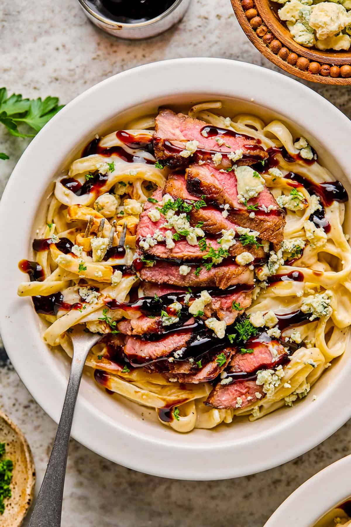 A bowl of creamy steak pasta topped with sliced steak, crumbled blue cheese, fresh herbs, and a drizzle of dark sauce, with a fork resting in the bowl.