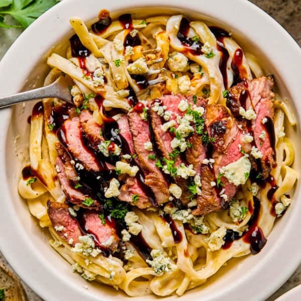 A bowl of creamy steak pasta topped with sliced grilled steak, crumbled blue cheese, chopped herbs, and a drizzle of dark balsamic glaze, with a fork resting in the bowl.