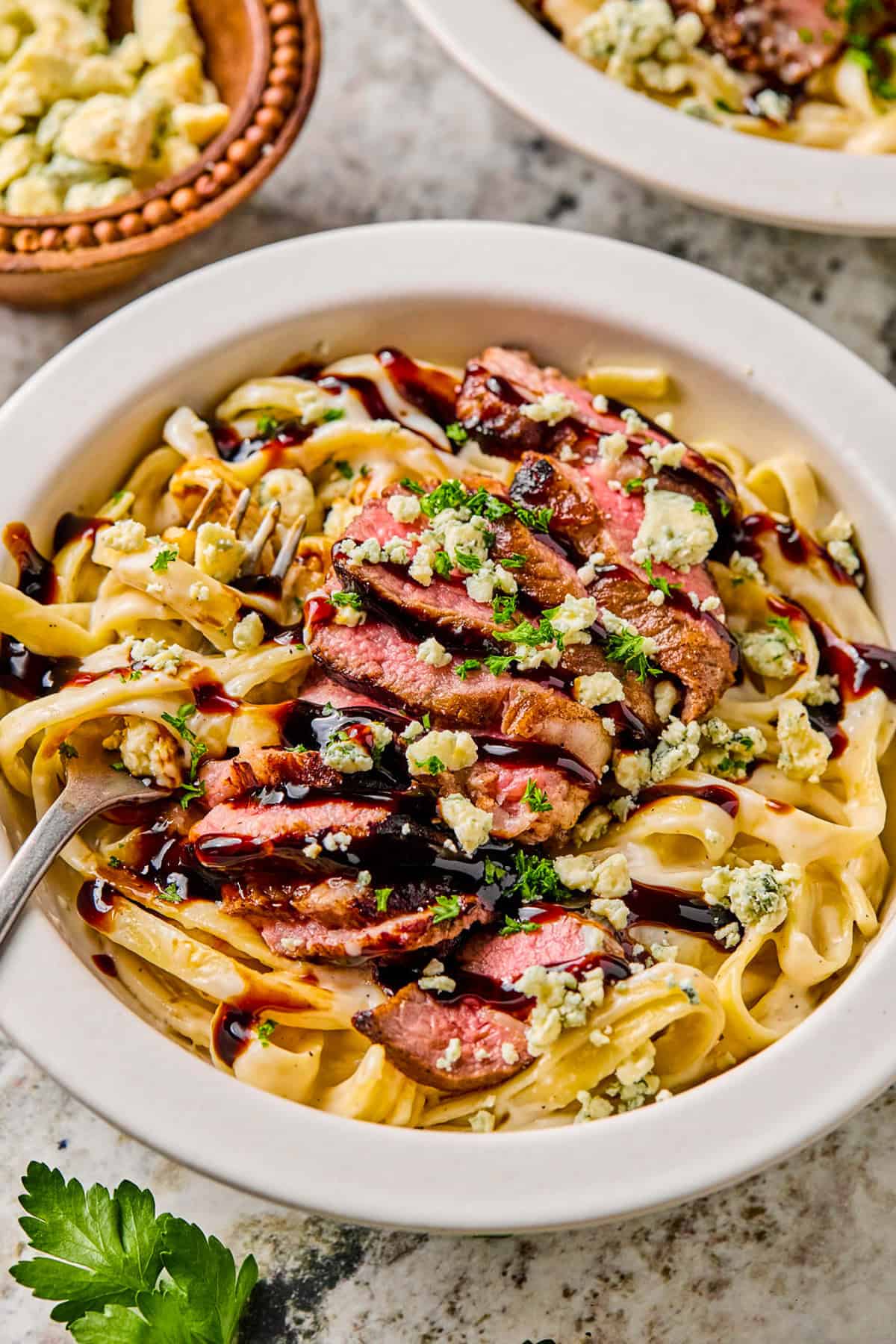 A bowl of steak pasta featuring fettuccine topped with sliced steak, crumbled blue cheese, chopped parsley, and a drizzle of dark sauce, with a fork in the bowl and a small dish of blue cheese in the background.