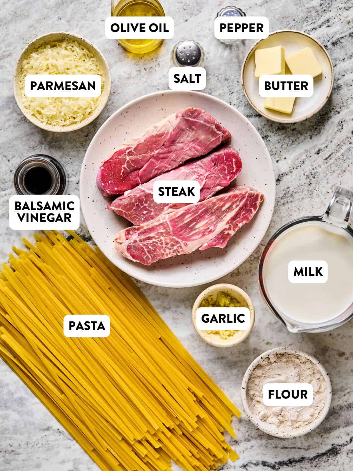 Top-down view of steak pasta ingredients on a marble surface, labeled: raw steak, uncooked pasta, grated parmesan, olive oil, salt, pepper, butter, milk, flour, minced garlic, and balsamic vinegar.