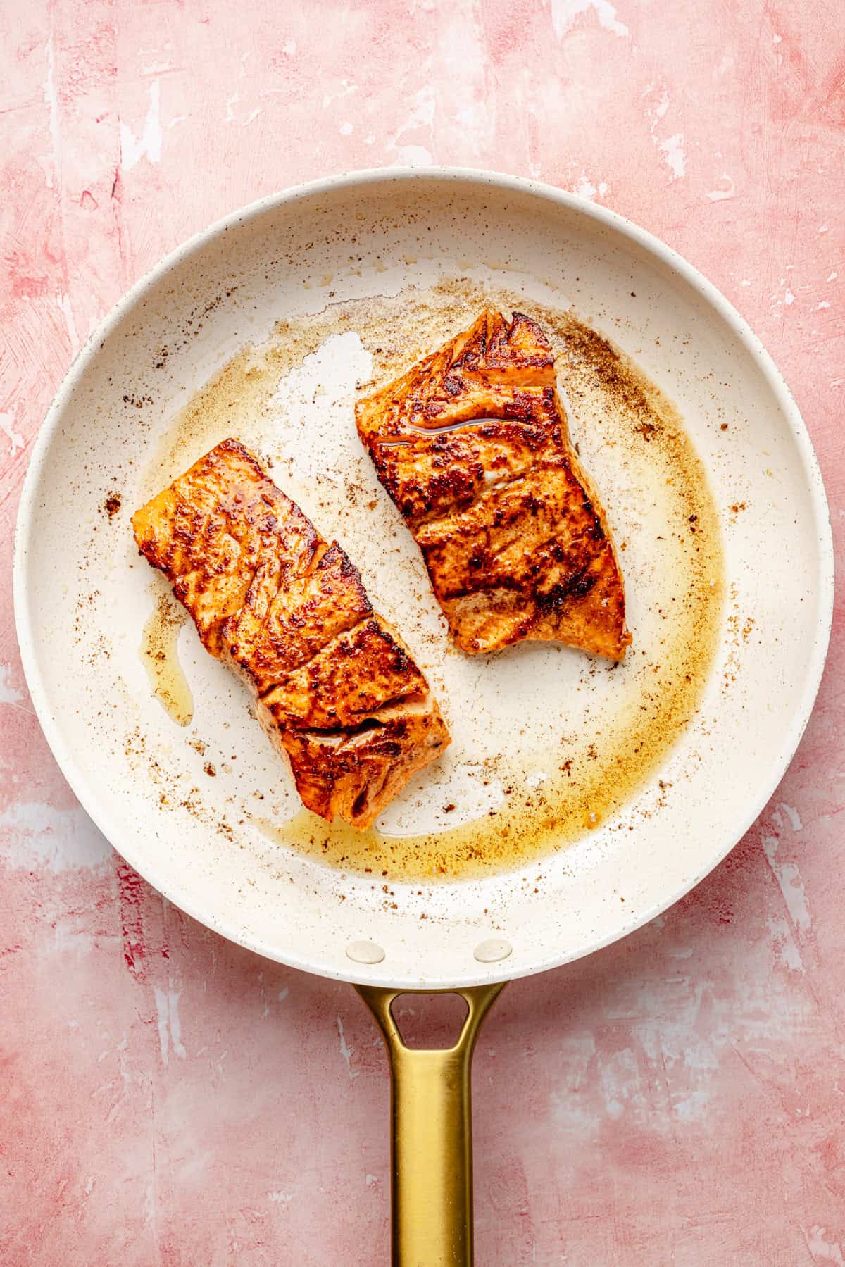 Two cooked salmon fillets with grill marks sit in a white frying pan with some oil, on a pink textured surface, ready to be flaked into a vibrant salmon pesto pasta.