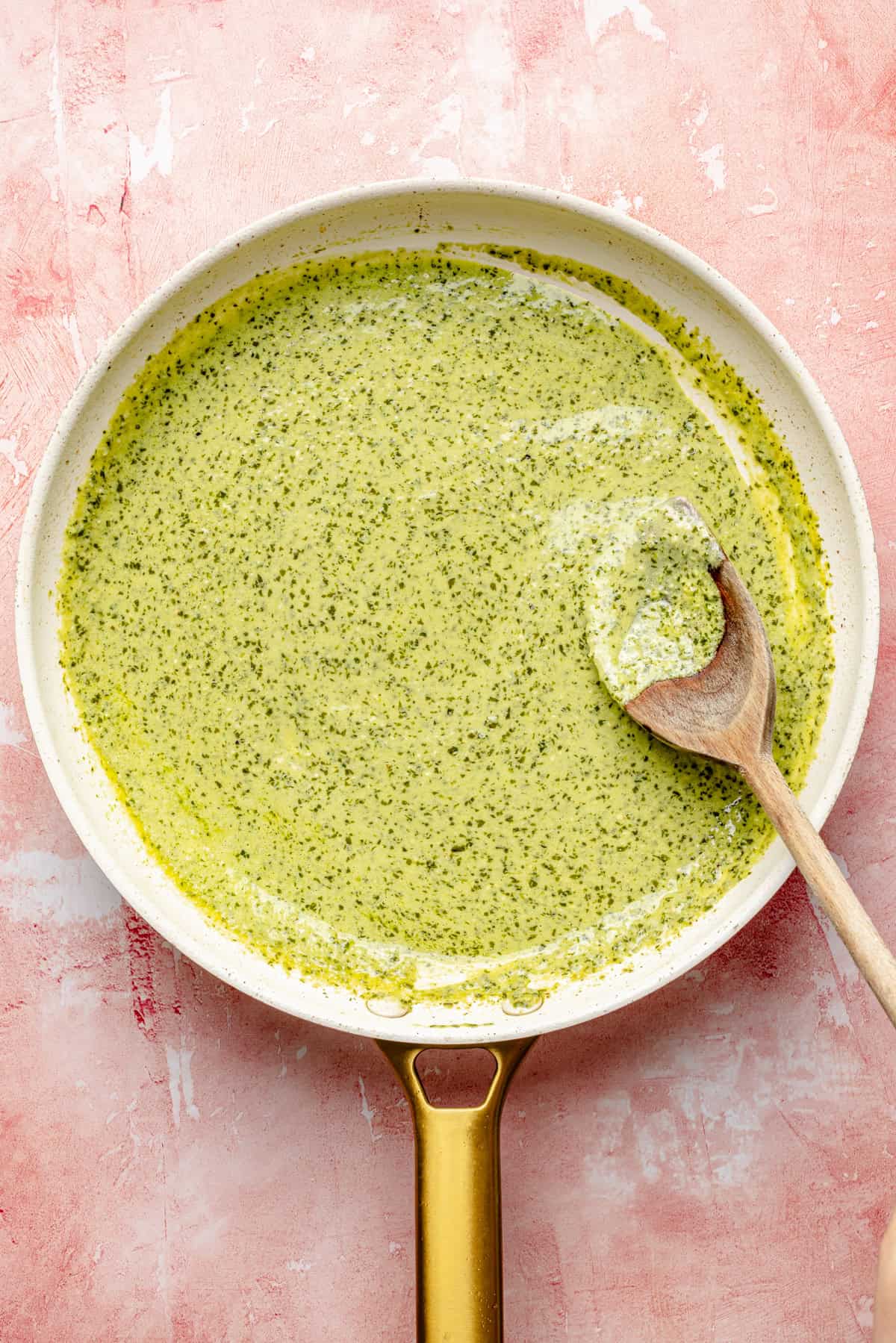A white skillet with green pesto cream sauce for salmon pesto pasta being stirred by a wooden spoon, set on a pink textured surface.
