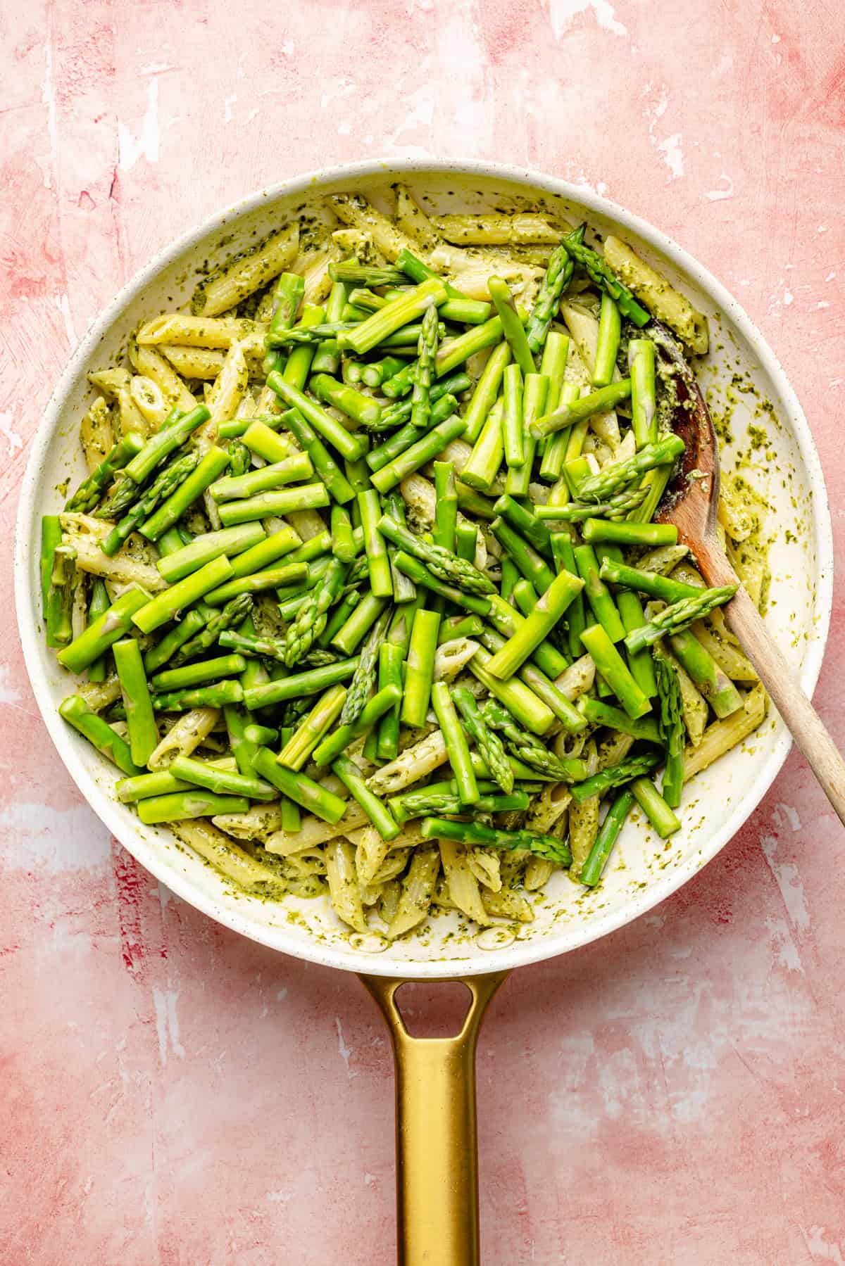 A skillet filled with salmon pesto pasta, featuring penne tossed in pesto sauce, tender pieces of bright green asparagus, and succulent flakes of salmon, with a wooden spoon resting inside—set against a pink textured background.