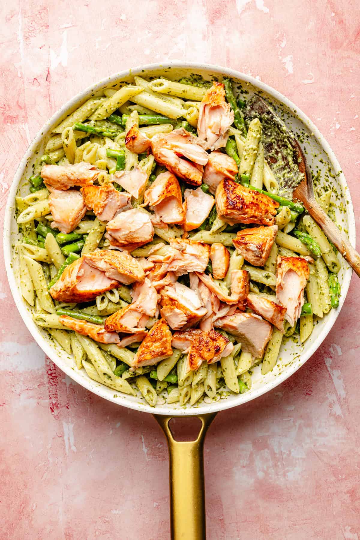 A gold-handled skillet filled with salmon pesto pasta—penne coated in vibrant green pesto sauce and topped with chunks of grilled salmon—sits on a pink textured surface, with a wooden spoon on the side.