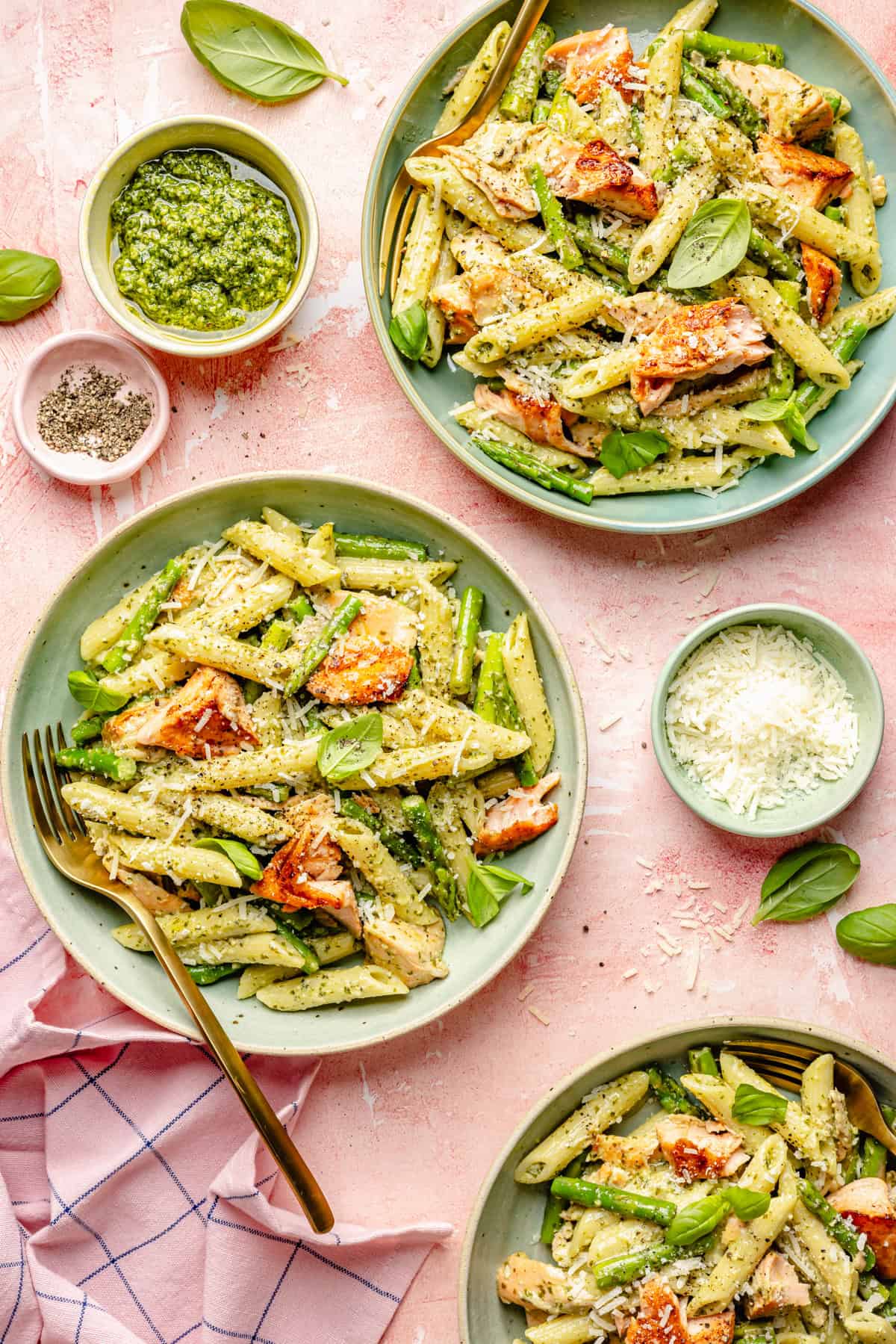 Three plates of penne pasta with pesto, seared salmon, and asparagus. Bowls of pesto, grated cheese, and pepper sit nearby. Basil leaves garnish the plates on a pink surface with a fork and a pink towel—perfect inspiration for your next salmon pesto pasta creation.