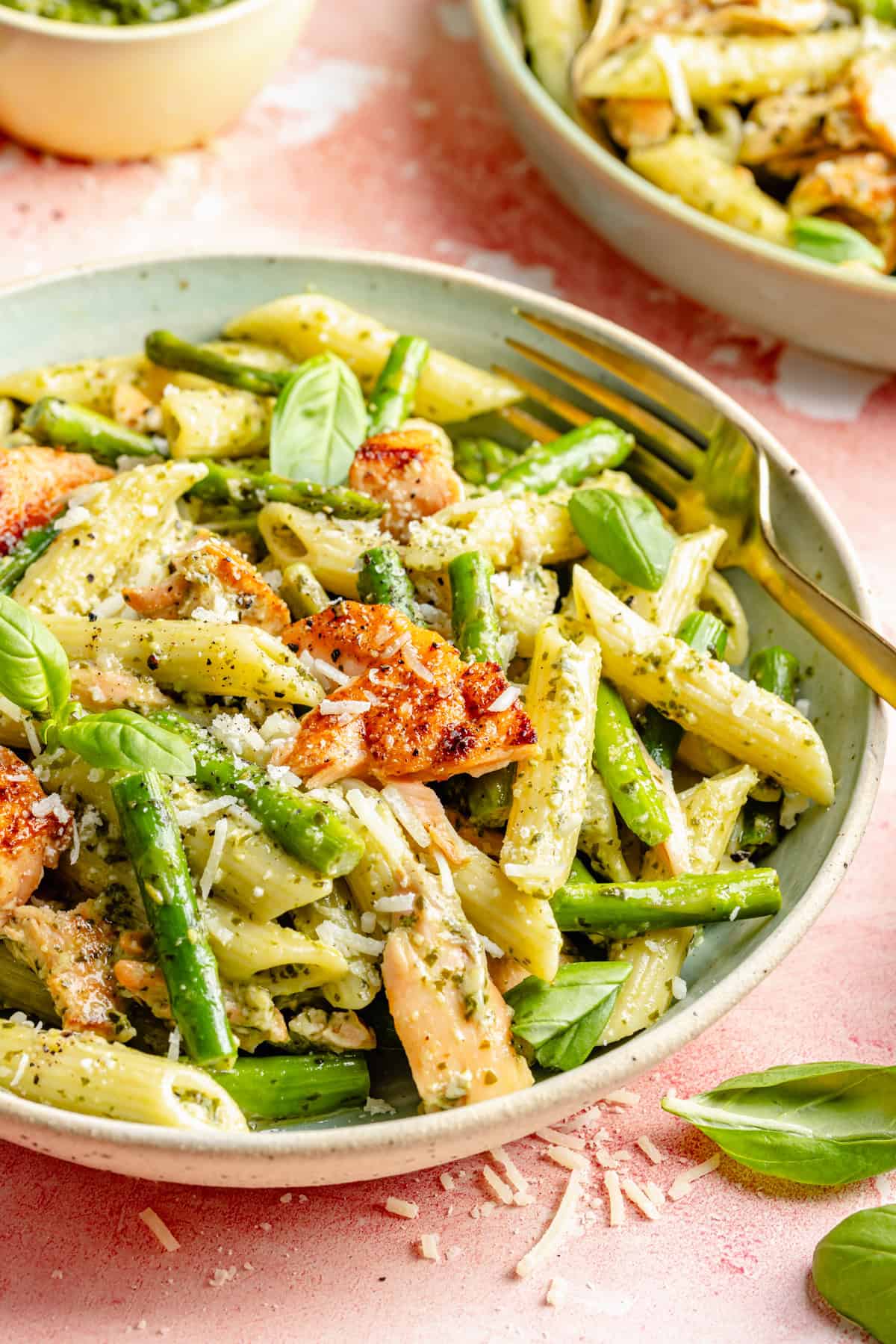 A bowl of penne pasta with asparagus, salmon pieces, fresh basil leaves, pesto sauce, and grated parmesan cheese—served salmon pesto pasta style—paired with a gold fork on a pink table.