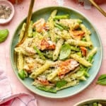 A plate of penne pasta with seared salmon, asparagus, pesto sauce, grated parmesan cheese, and fresh basil leaves—reminiscent of a classic salmon pesto pasta—served with a gold fork.