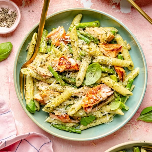 A plate of penne pasta with seared salmon, asparagus, pesto sauce, grated parmesan cheese, and fresh basil leaves—reminiscent of a classic salmon pesto pasta—served with a gold fork.