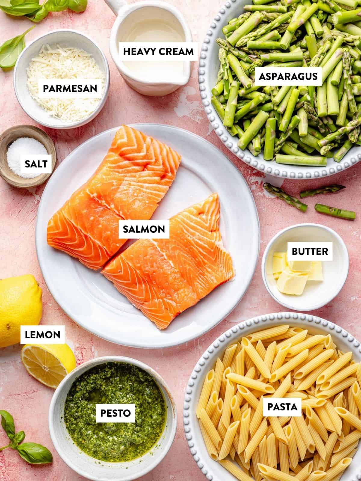 A flat lay of labeled ingredients for salmon pesto pasta: salmon, asparagus, pasta, pesto, lemon, salt, butter, heavy cream, and grated parmesan arranged in bowls and plates over a pink background.