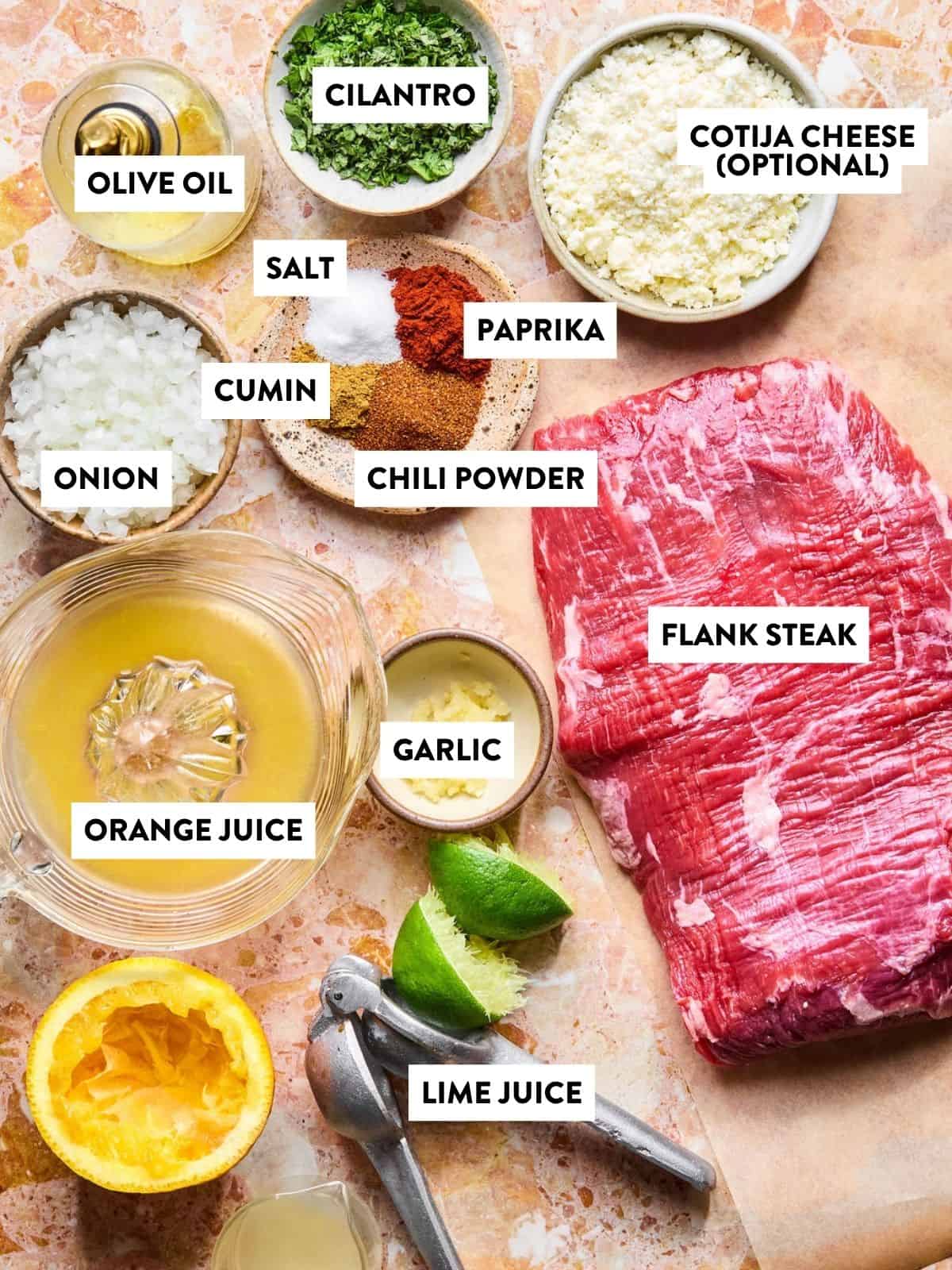 Raw flank steak on a cutting board, ready for carne asada tacos, surrounded by labeled ingredients: chopped cilantro, Cotija cheese (optional), olive oil, salt, paprika, cumin, chili powder, chopped onion, orange juice, minced garlic, and lime juice.