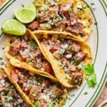 Three grilled carne asada tacos topped with chopped onions, cilantro, and crumbled cheese are served on a plate with lime wedges and a fresh cilantro garnish.