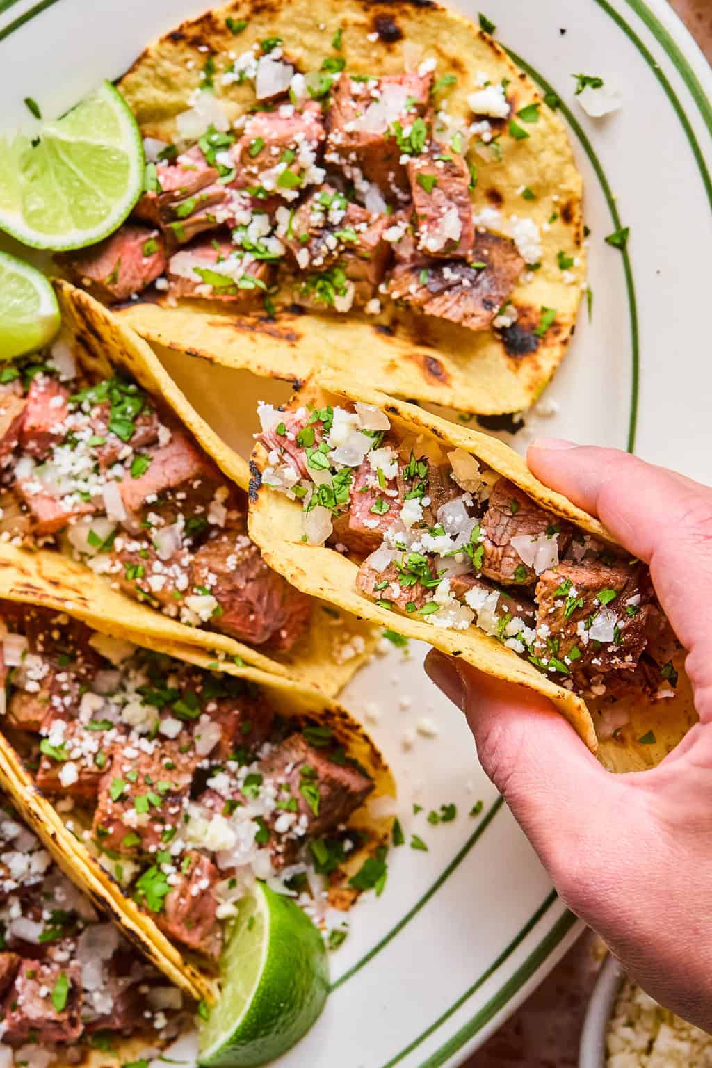 A hand holding a carne asada taco topped with chopped onions, cilantro, and cheese rests on a plate with more carne asada tacos and lime wedges.