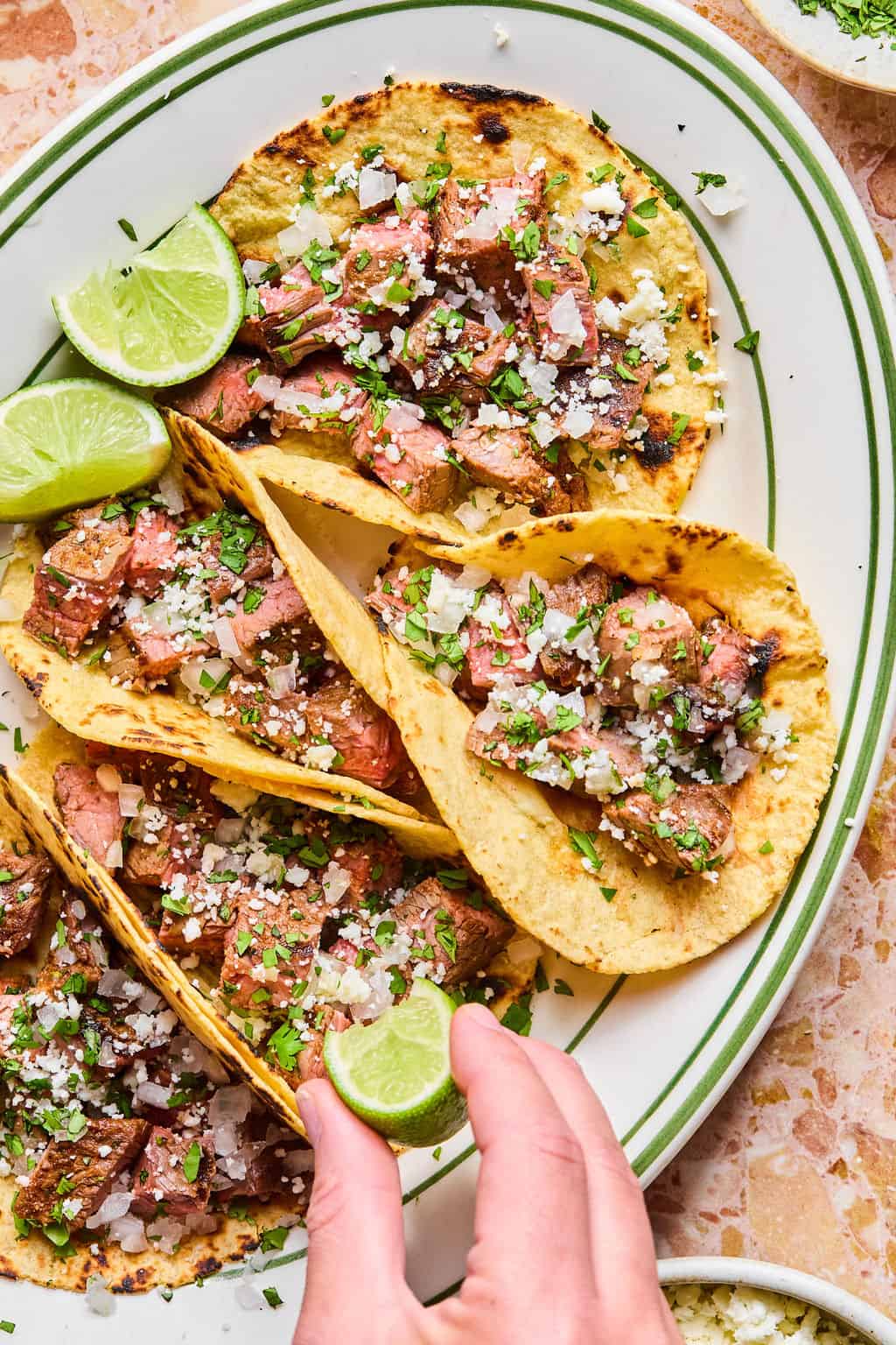 A hand squeezes a lime wedge over carne asada tacos topped with chopped cilantro and crumbled cheese, served on a white plate with extra lime slices.