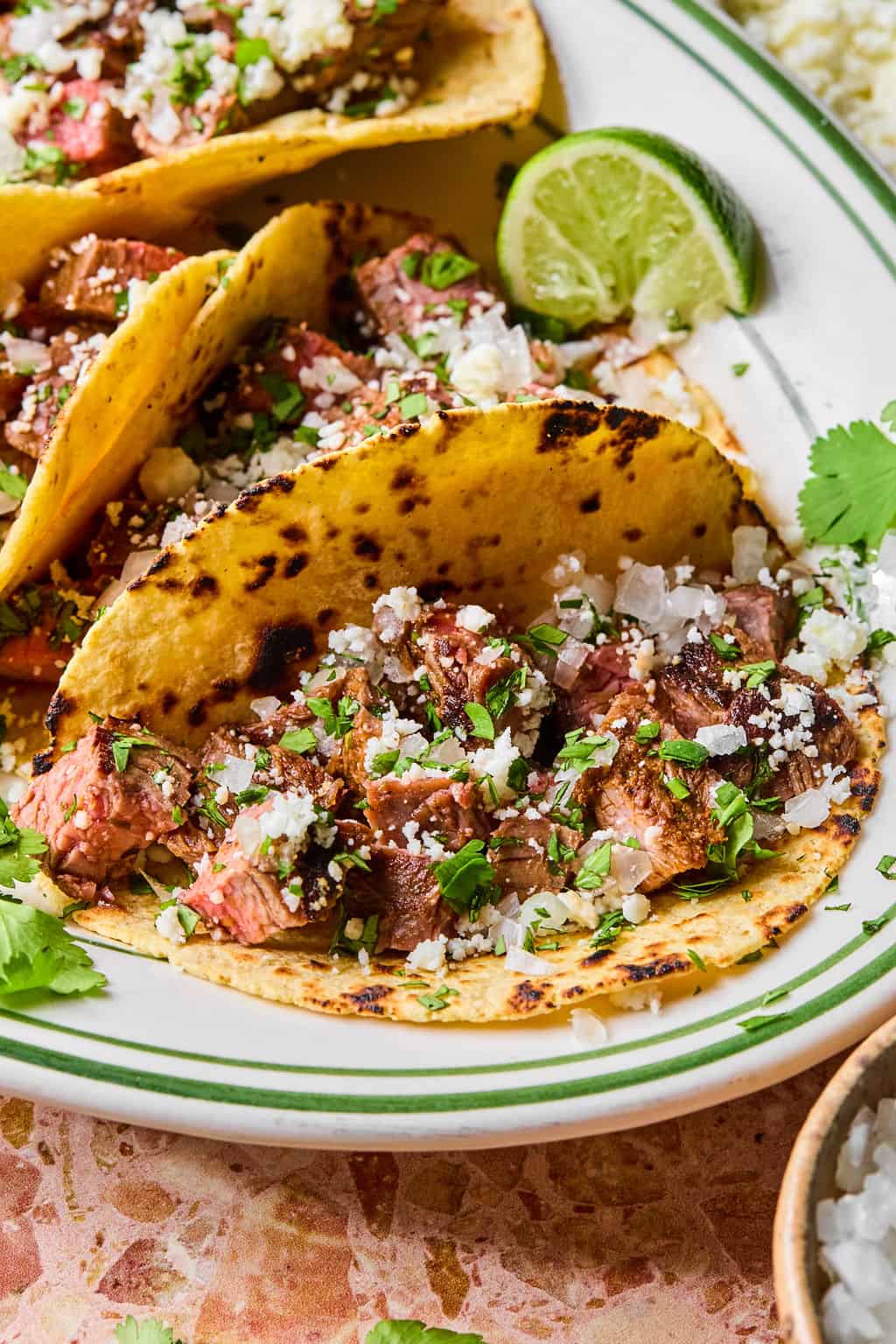 Two grilled carne asada tacos topped with chopped onions, cilantro, and crumbled cheese sit on a white plate with green trim, garnished with lime wedges and fresh cilantro.