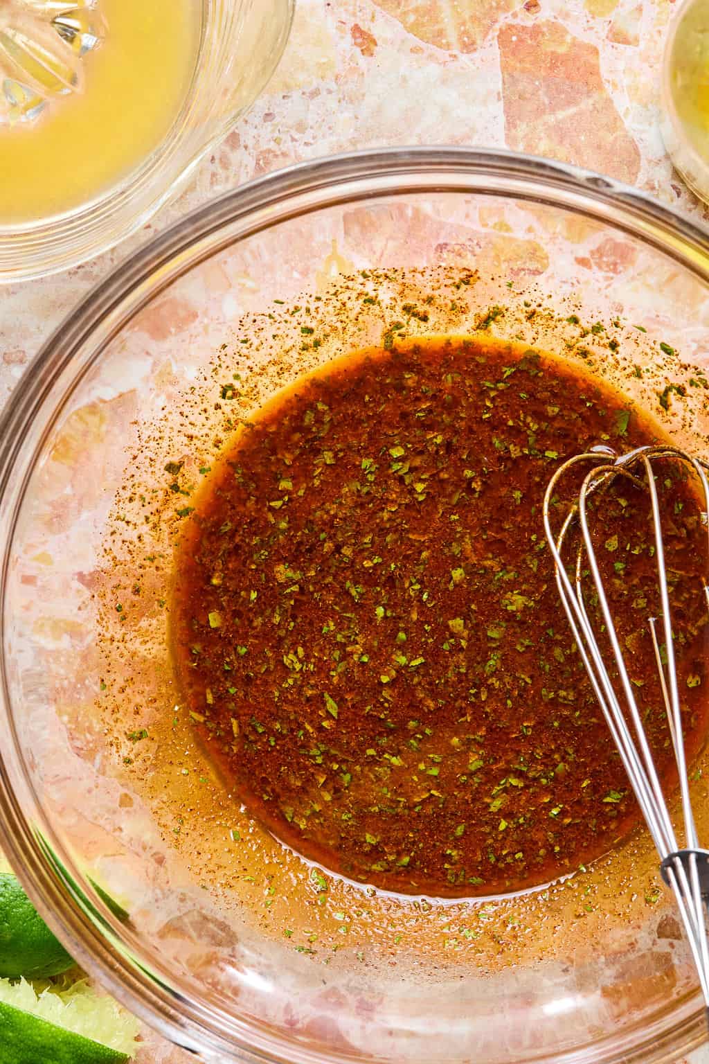 A glass bowl filled with a reddish-brown marinade, perfect for seasoning carne asada tacos, is sprinkled with green herbs and sits on a marble countertop. A small metal whisk rests inside the bowl, with lime wedges visible in the corner.