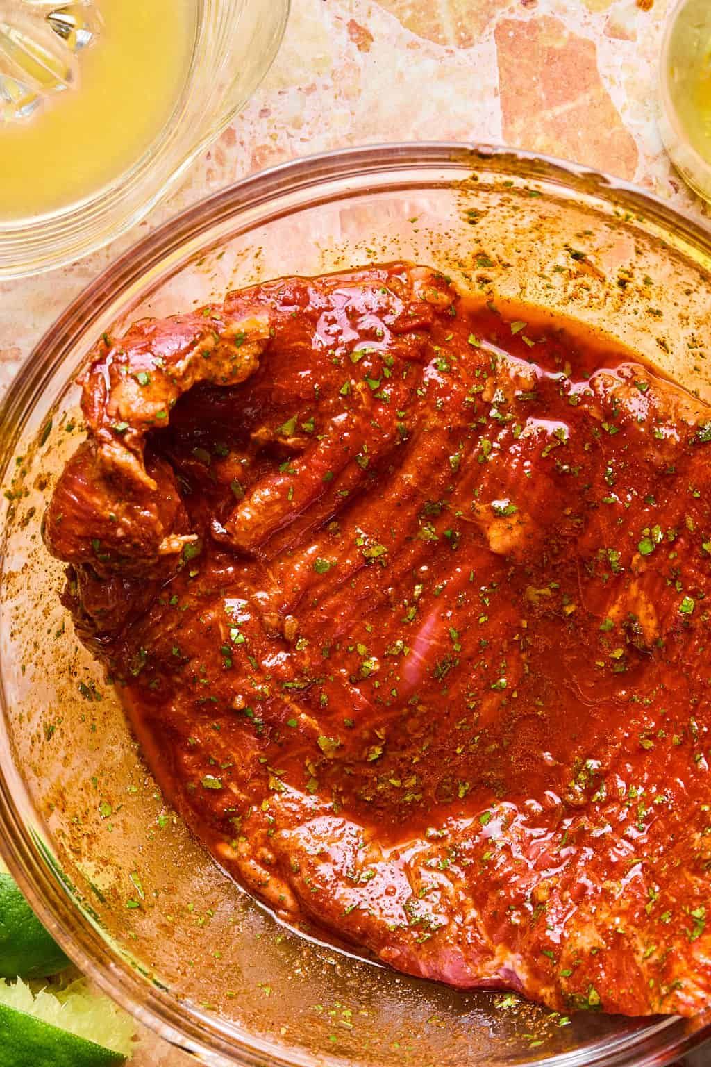 A raw piece of flank steak coated in a reddosj carme asada marinade with herbs sits in a clear glass bowl on a stone countertop, ready to be transformed into delicious carne asada tacos. Green lime wedges and parts of two drink glasses are visible at the edges.