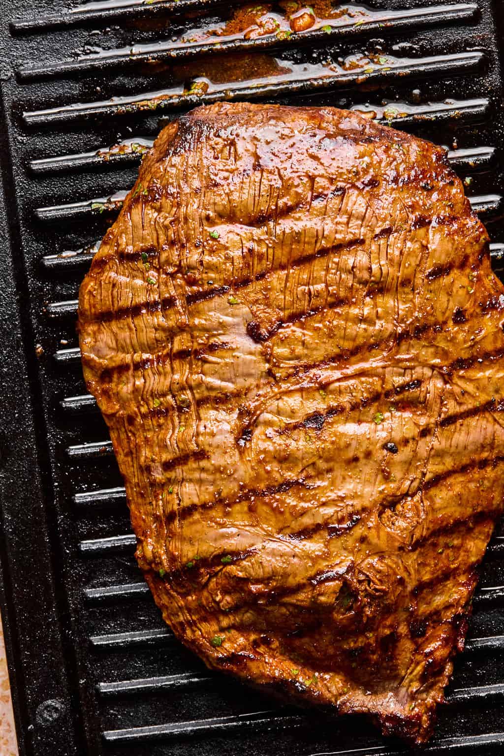 Grilled marinated flank steak on a ridged grill pan, showing sear marks and a glossy, seasoned surface—perfect for carne asada tacos.