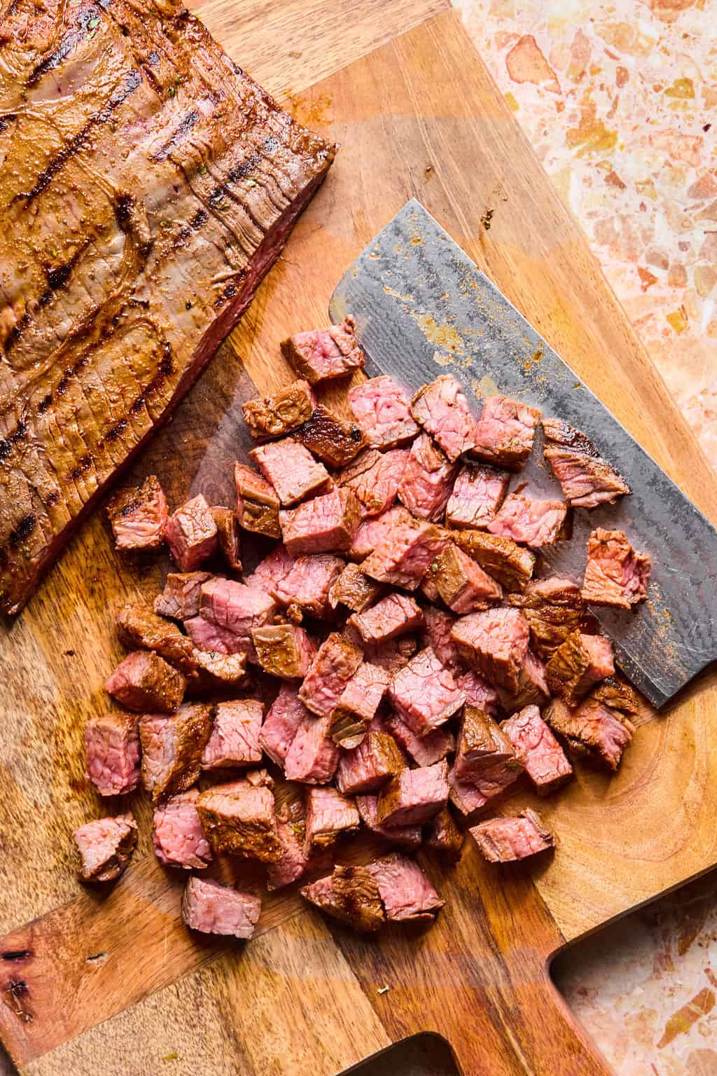 Sliced grilled steak is chopped into small cubes on a wooden cutting board, ready to fill carne asada tacos. A large knife rests nearby, and the juicy steak appears cooked to medium doneness.