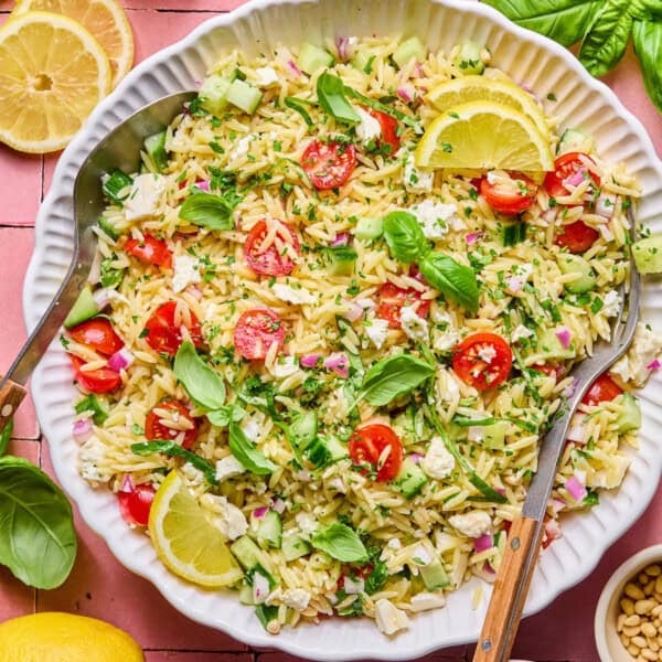 A bowl of lemon basil orzo salad with cherry tomatoes, cucumber, red onion, feta cheese, fresh basil, and lemon slices, served with two wooden-handled salad servers.