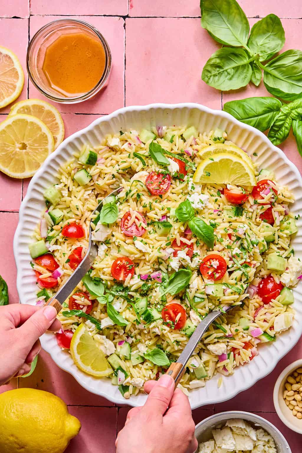 A person tosses a vibrant lemon basil orzo salad with cherry tomatoes, cucumber, red onion, feta, and lemon slices. Nearby, a jar of dressing, lemon halves, basil leaves, and pine nuts sit on a pink tiled surface.