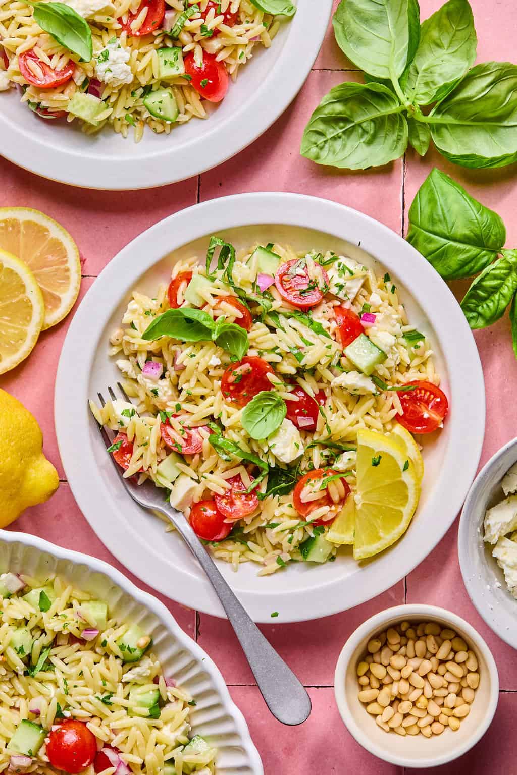 Overhead view of a lemon basil orzo salad with cherry tomatoes, cucumbers, feta cheese, red onions, and fresh basil, garnished with lemon wedges. Surrounding are fresh basil leaves, lemon slices, pine nuts, and additional salad.