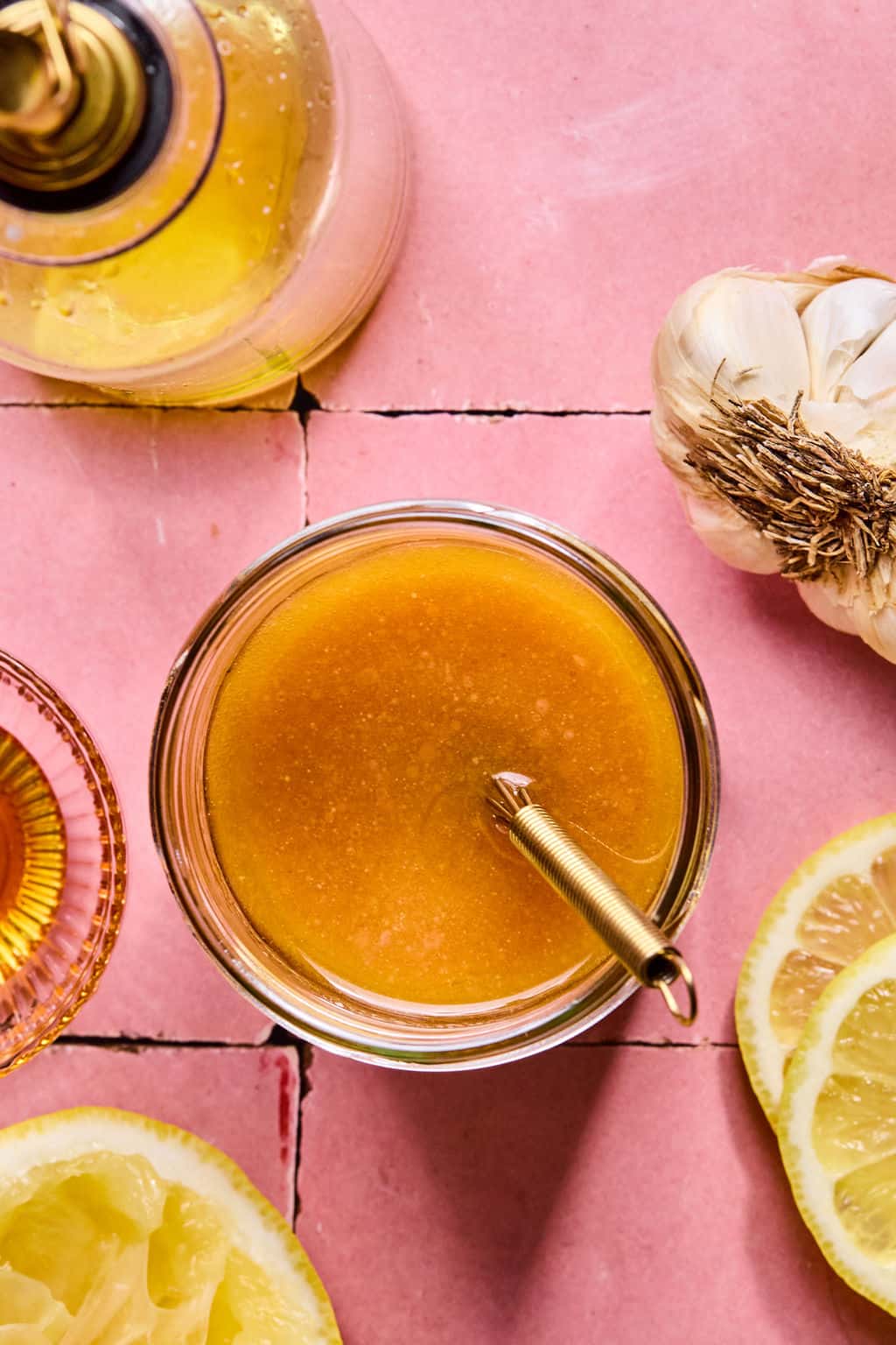 A glass jar filled with honey mustard dressing, perfect for drizzling over lemon basil orzo salad, sits with a spoon inside, surrounded by a lemon half, garlic head, oil bottle, and small bowl on a pink tiled surface.