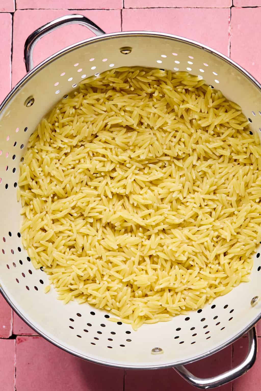 Cooked orzo pasta, perfect for a lemon basil orzo salad, is draining in a metal colander set on a pink tiled surface. The small, rice-shaped pasta fills most of the colander, with steam holes visible around its edges.