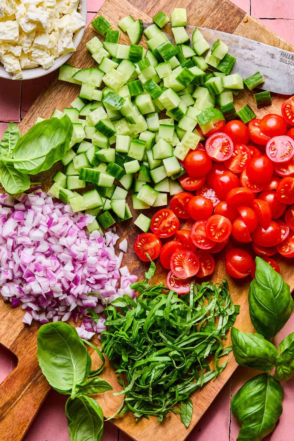 A wooden cutting board with chopped cucumber, halved cherry tomatoes, diced red onion, shredded basil, and whole basil leaves—perfect prep for a vibrant lemon basil orzo salad. A bowl of feta cheese and a knife are also visible.