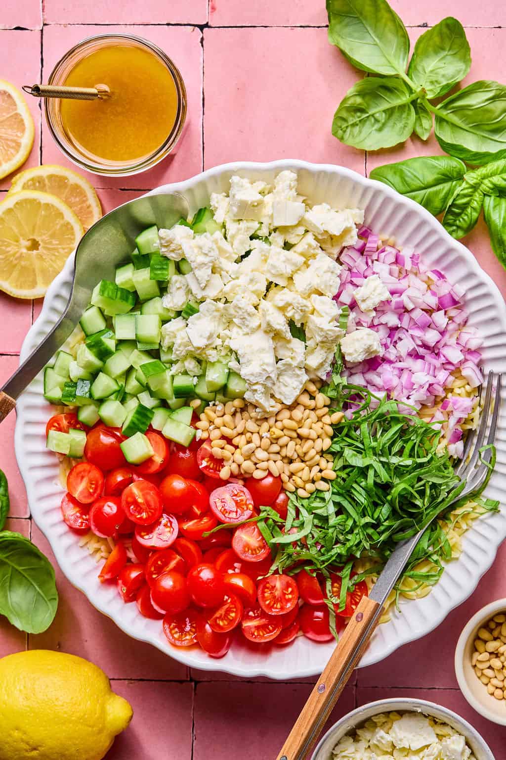 A large white bowl filled with chopped cucumbers, cherry tomatoes, red onion, feta cheese, pine nuts, and shredded basil—this vibrant spread is perfect for a lemon basil orzo salad, surrounded by fresh basil leaves, lemon, and vinaigrette on a pink tiled surface.