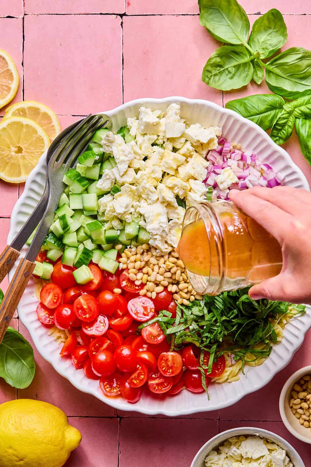 A hand pours dressing from a jar over a vibrant lemon basil orzo salad with cherry tomatoes, cucumber, red onion, feta, pine nuts, and basil in a white bowl. Utensils, lemon slices, and basil leaves are arranged around the bowl on a pink surface.