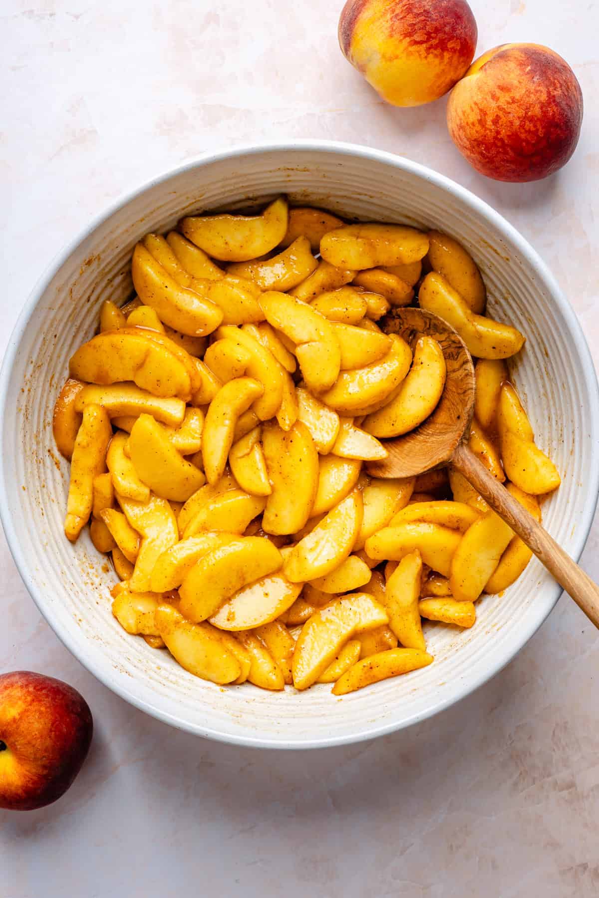 Sliced peaches coated in spices and sugar in a white bowl with a wooden spoon, ready to be transformed into a delicious gluten free peach cobbler. Whole peaches are placed around the bowl on a light surface.